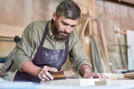Portrait of mature bearded carpenter sanding wooden part in modern workshop, copy space